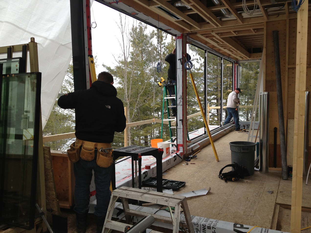 Installation in progress at a Muskoka cottage with Reynaers Aluminum MasterLine 8 window systems, showing installers fitting large fixed windows overlooking the forest.