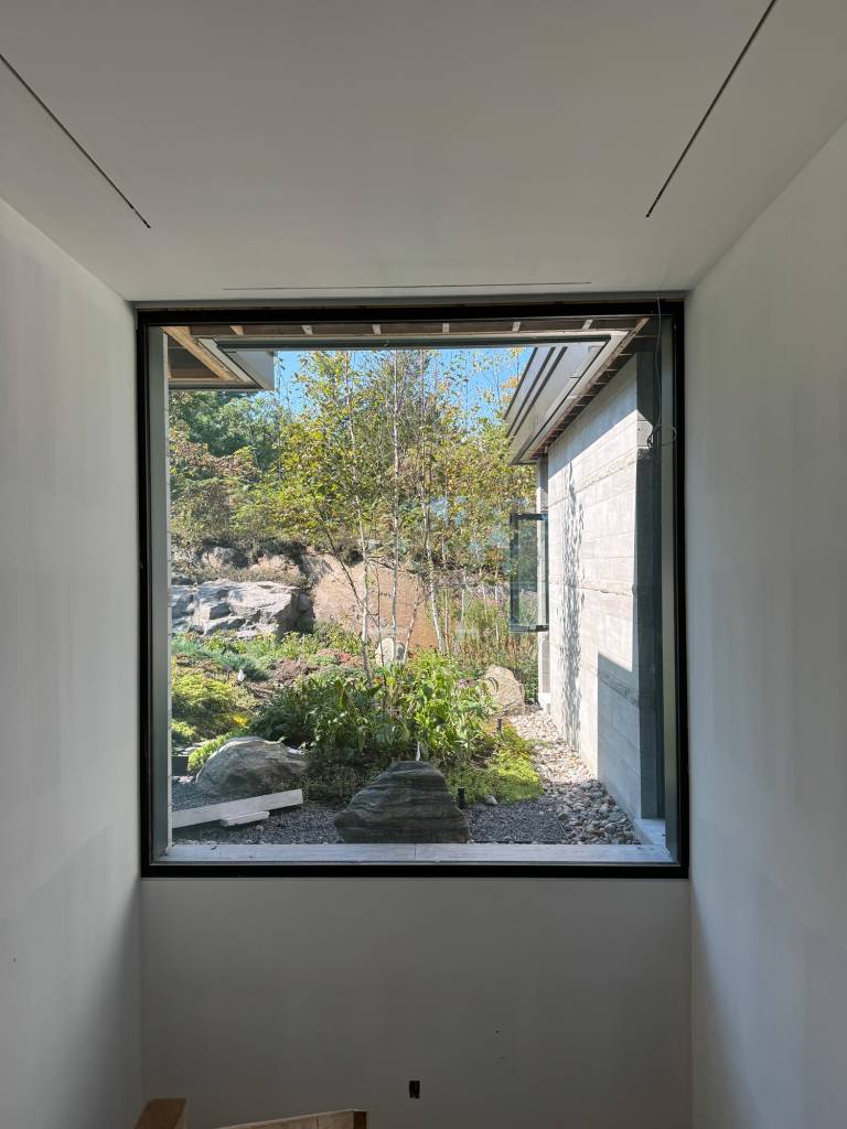 Interior view of a stairwell at a St. Lawrence River home featuring a large Reynaers Aluminum window, framing landscaped stone and garden views with modern glazing.