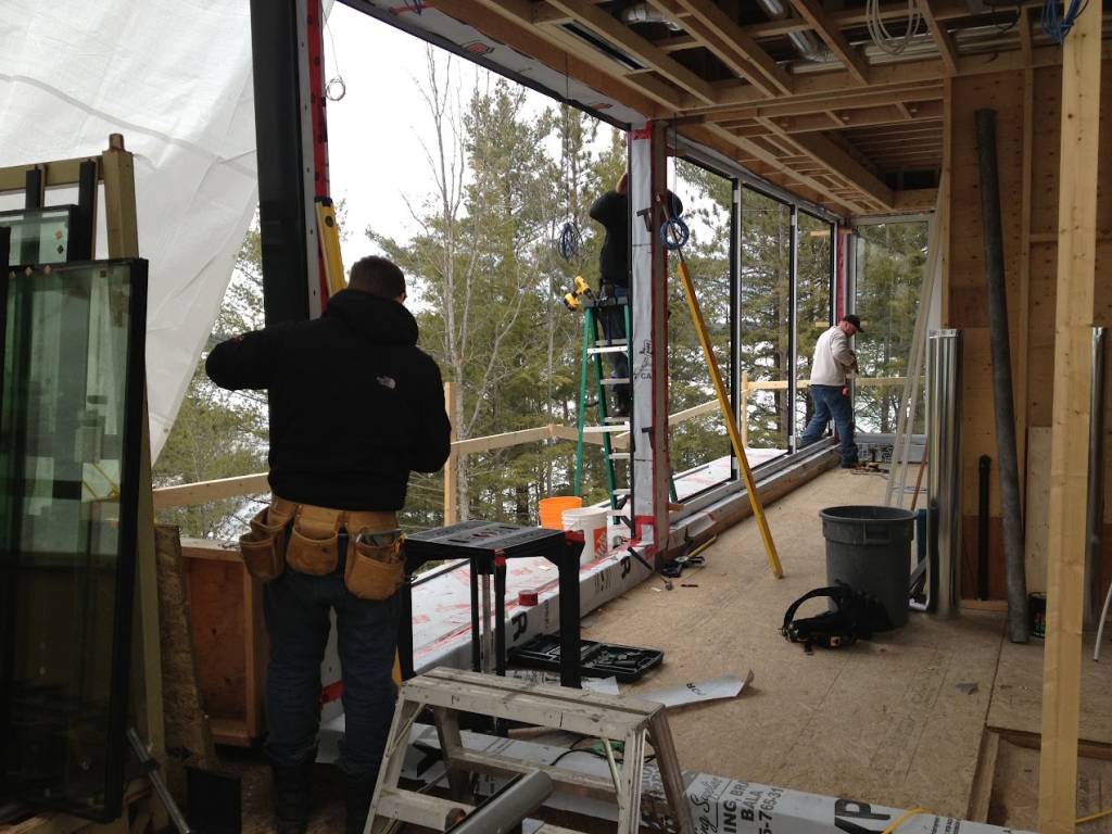 Installation in progress at a Muskoka cottage with Reynaers Aluminum MasterLine 8 window systems, showing installers fitting large fixed windows overlooking the forest.