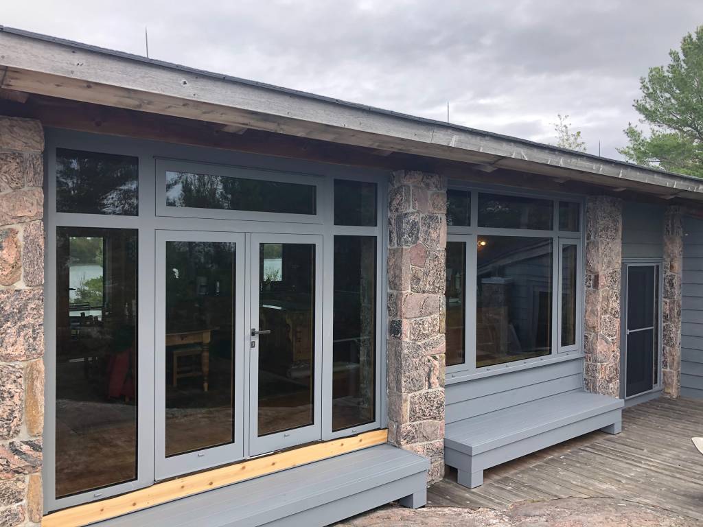 Exterior view of a Georgian Bay island cottage with Reynaers CS77 dual-colour French doors and windows framed in stone and wood cladding.