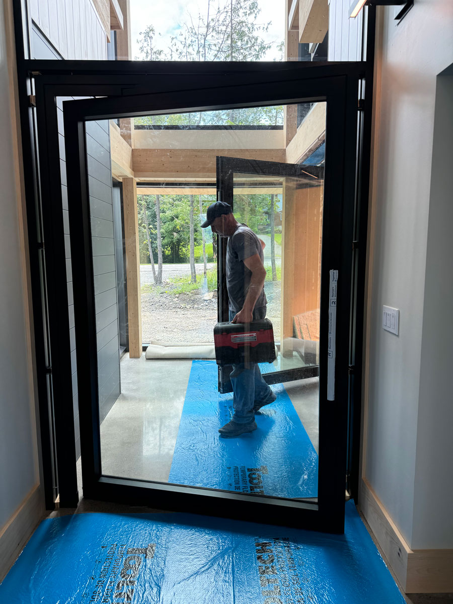 Interior hall view of a 4.5' x 7.5' Reynaers MasterLine 8 black pivot door set in curtain wall framing with glass above, Ottawa River home.