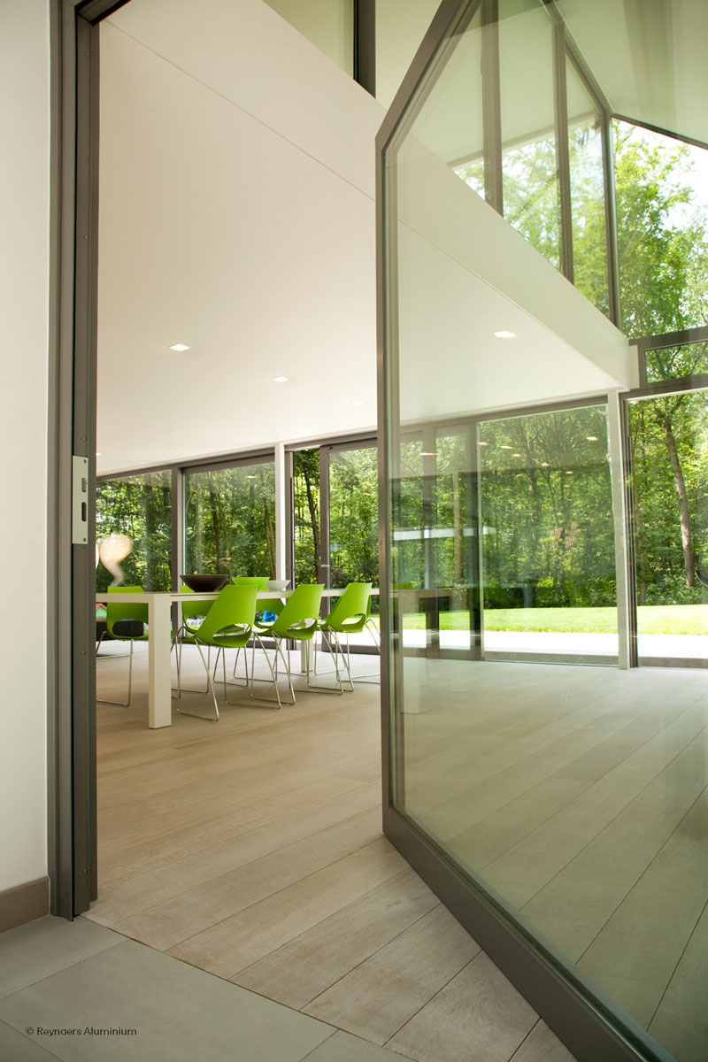 Reynaers MasterLine 8 pivot door partially open in a modern private house in Loppem, showing glass walls and interior dining area.